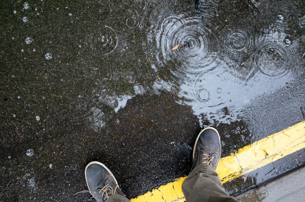 Rainy weather. Male legs in sneakers or boots walking through the rain puddle on the asphalt road, top view. Film grain photo.