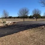 freshly paved asphalt driveway bordered by grass and trees