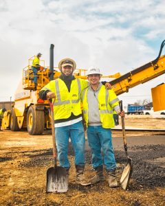 2 men in construction vests holding shovels in front of an excavator