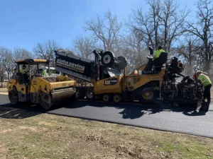 Fresh asphalt overlay being installed with paving machine, dump truck, and roller during roadway resurfacing project.