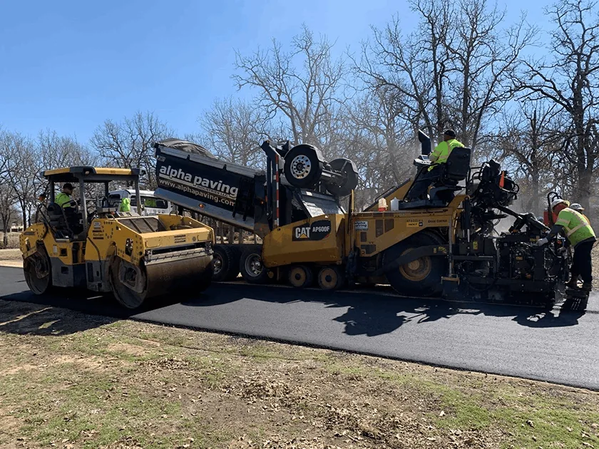 Fresh asphalt overlay being installed with paving machine, dump truck, and roller during roadway resurfacing project.