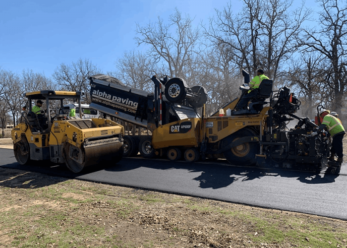 Fresh asphalt overlay being installed with paving machine, dump truck, and roller during roadway resurfacing project.